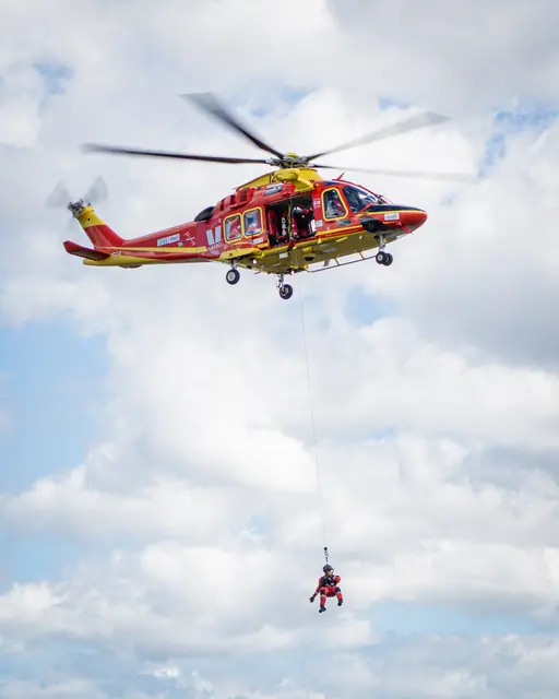 Rescue helicopter winching a crew member beneath the aircraft against a cloudy sky