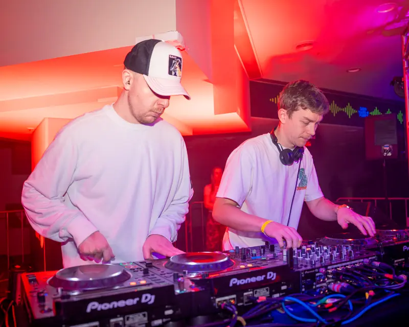 Two DJs performing together under pink and blue club lighting